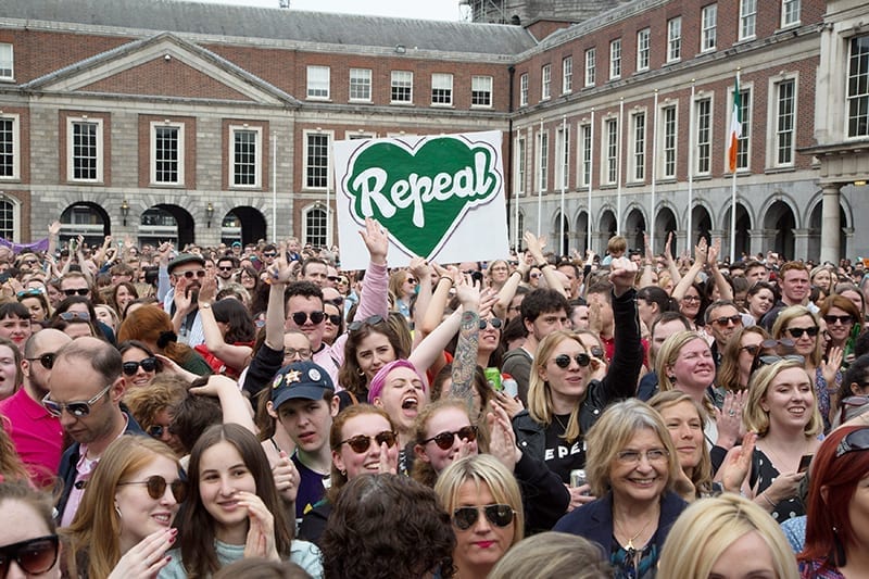 A crowd celebrating the repeal of the 8th Amendment in Ireland. In the center a hand holds up a sign saying "Repeal" in a green heart.