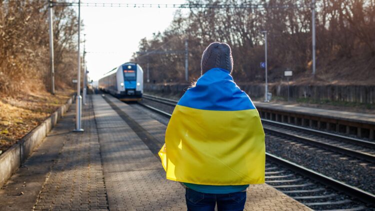 A woman wrapped in the Ukrainian flag stands at a platform watching a train arrive.