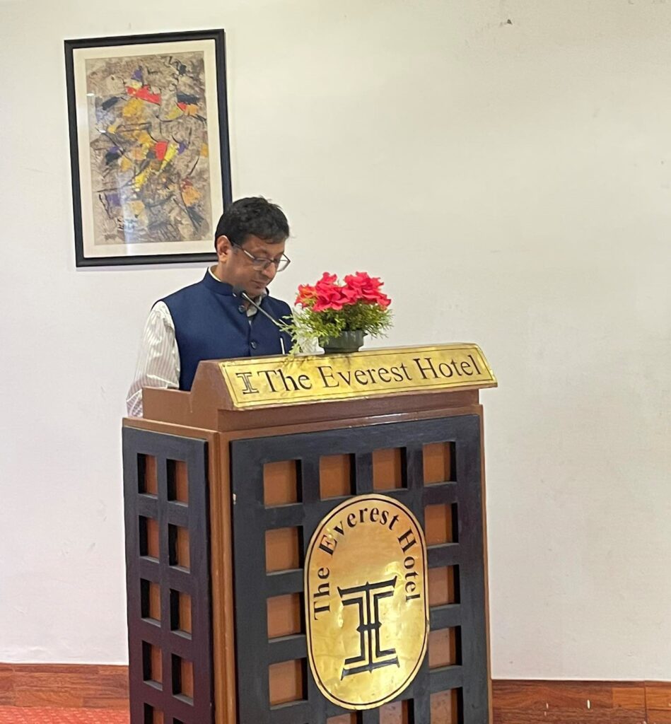 A man speaks at a podium with several signs on it reading "The Everest Hotel." A vase of red flowers sits on top of the podium and an abstract painting hangs in the background.