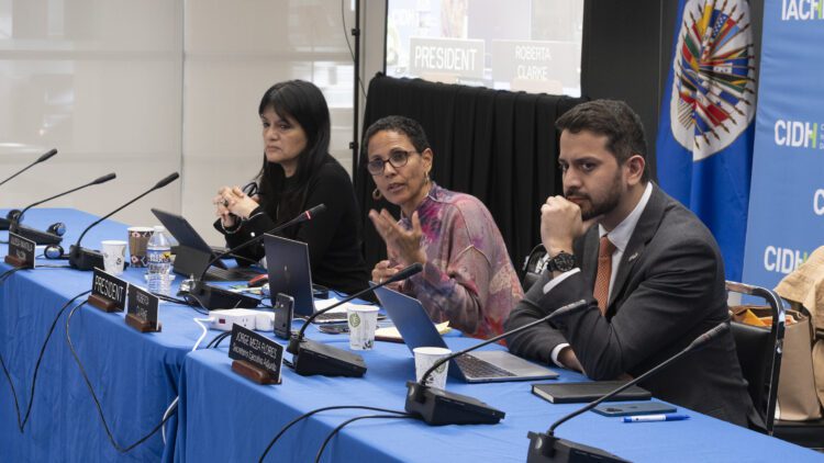 Three participants in the IACHR hearing sit at a table behind laptops and microphones. One woman is speaking and gesturing toward the viewer.
