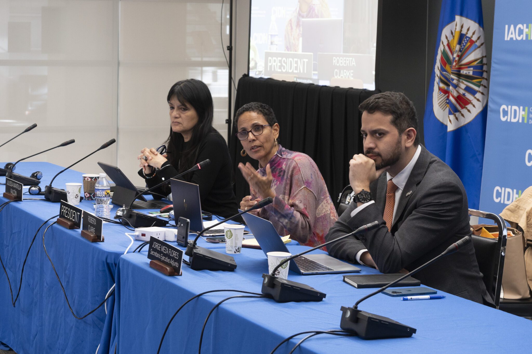 Three participants in the IACHR hearing sit at a table behind laptops and microphones. One woman is speaking and gesturing toward the viewer.