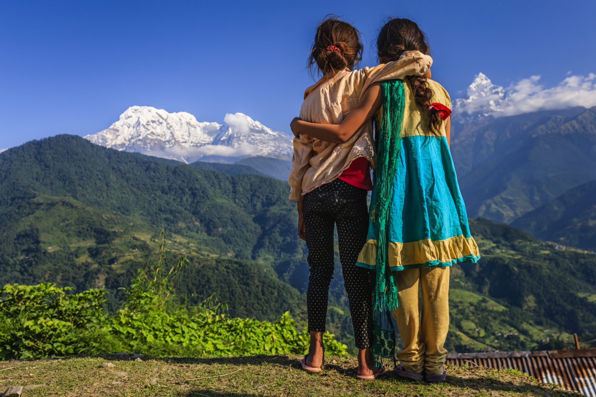 Two young girls stand with their arms around each other and their backs to the viewer, looking out at a mountain range.