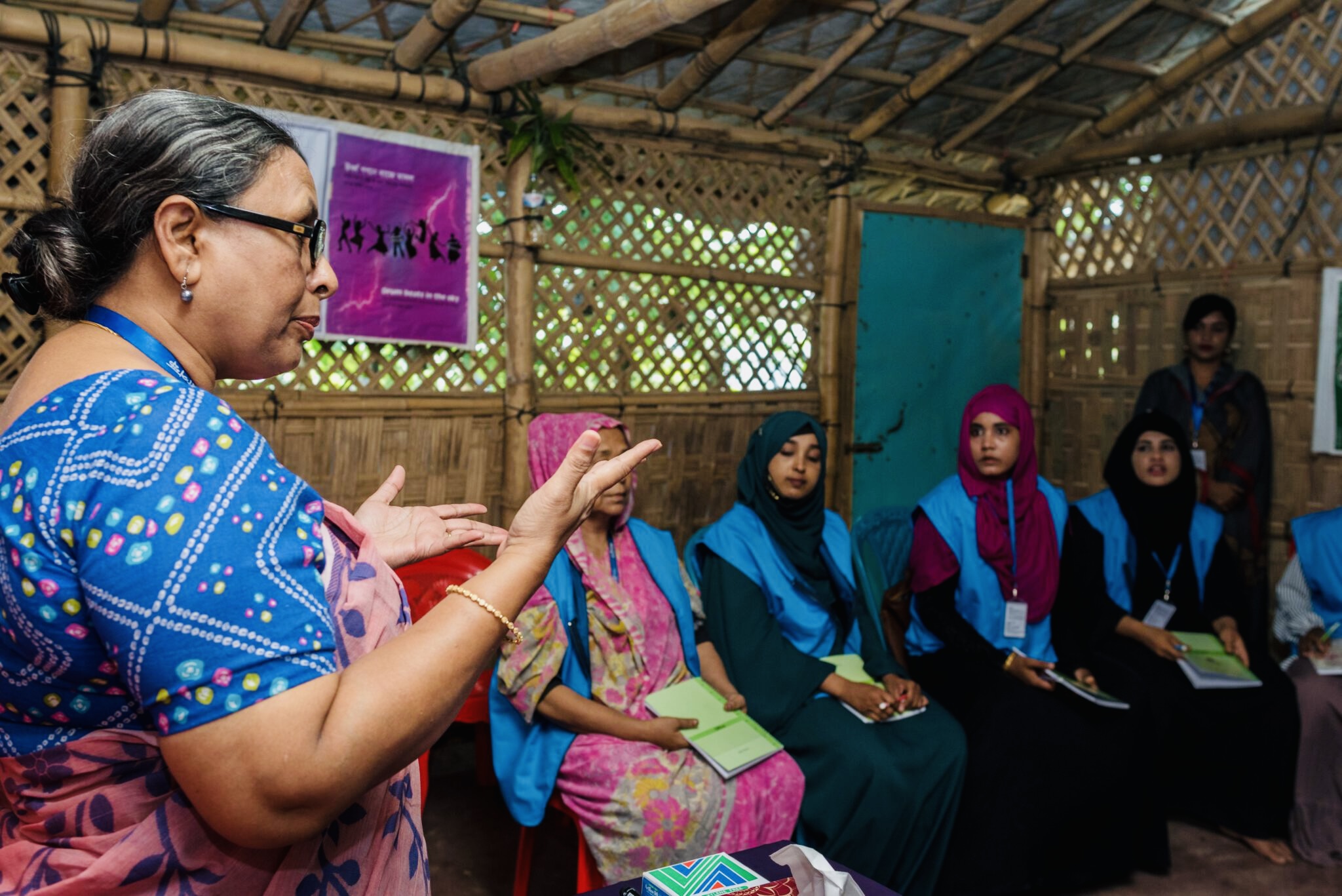 A woman stands in a wooden structure, speaking to several other women seated in a row nearby. Bangladesh Program to Improve Refugees' Reproductive Health Services