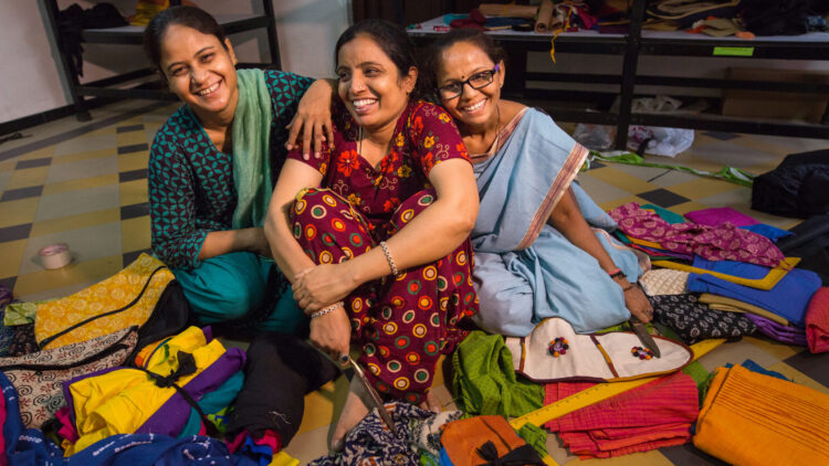 Three Indian women sit on a floor, smiling with their arms around each other.