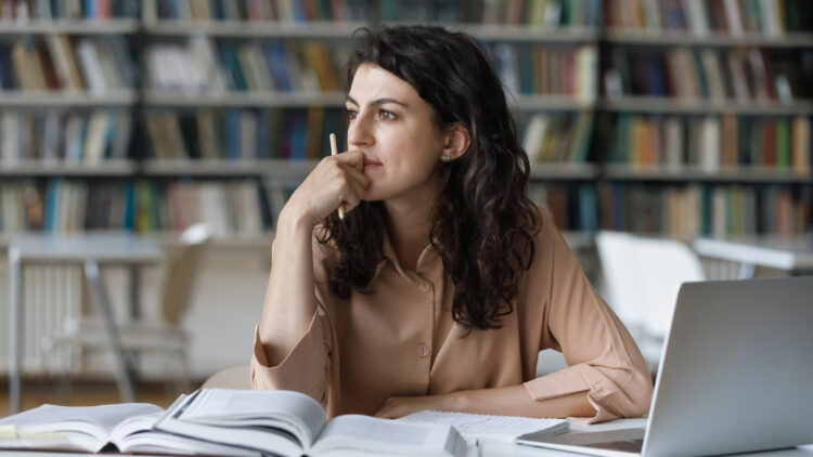 Woman / student seated in a library with her laptop and textbooks, looking pensively into the distance.
