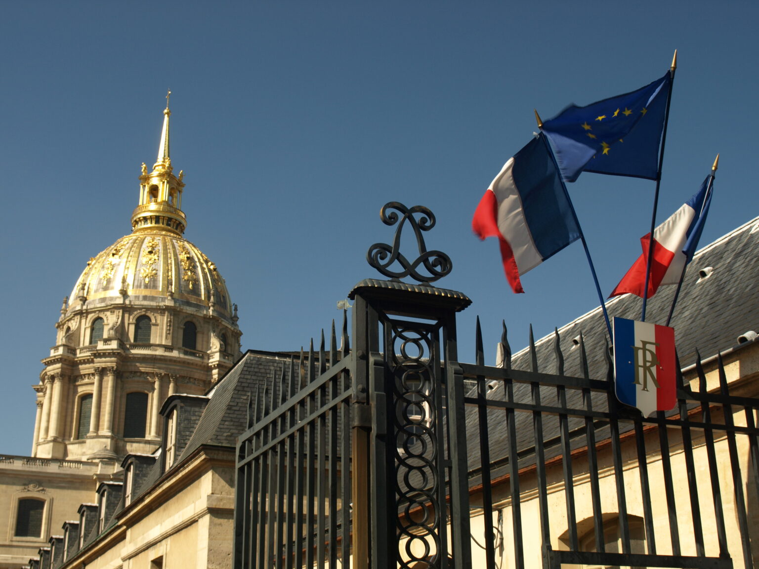 French and European Union flags waving outside the Dome des Invalides in Paris under a clear blue sky.